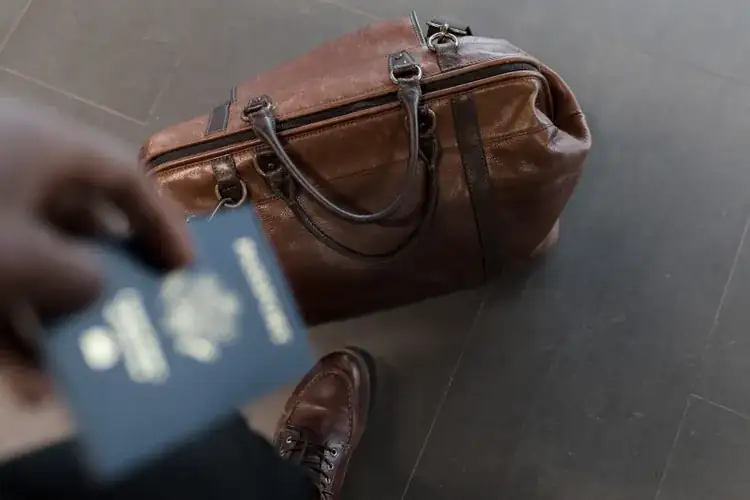 An orange suitcase covered in travel stickers standing on beach sand with the blue ocean behind it