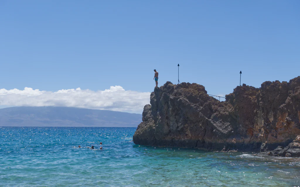 A cliff diver stands atop Black Rock at Kaanapali Beach Maui above turquoise water, with swimmers below and the island of Lanai across the channel
