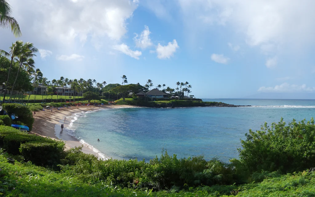The gold-sand crescent of Napili Bay Maui seen from above, ringed by palms and low-rise resorts with turquoise water and a rocky point