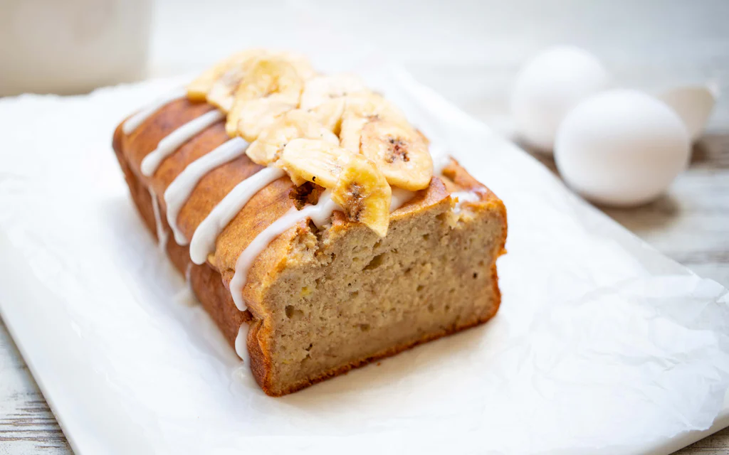 A warm golden banana bread loaf topped with banana chips on white parchment